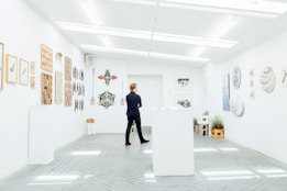 A person stands in a modern art gallery, surrounded by artwork hung on white walls. The gallery features various art pieces including paintings and sculptures. The lighting is bright and evenly distributed, creating a clean and open atmosphere. Plants add a touch of greenery to the otherwise minimalistic space.