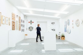 A person stands in a modern art gallery, surrounded by artwork hung on white walls. The gallery features various art pieces including paintings and sculptures. The lighting is bright and evenly distributed, creating a clean and open atmosphere. Plants add a touch of greenery to the otherwise minimalistic space.