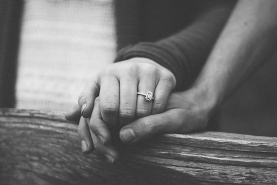 A black and white image showing a bride's delicate hands gently holding a groom's hand, with soft shadows casting depth.