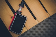 A vaping device with a sleek black body and red accents lies on a wooden surface with a light brown color. The vaping device has a transparent tank and is labeled with the brand 'SMOK'. The surface on which it rests appears to be part of a wooden pallet.