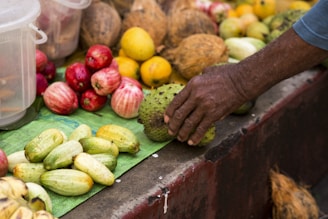 A display of various tropical fruits includes green and yellow cucumbers, red apples, yellow lemons, and coconuts. A hand is selecting one of the fruits, indicating an interaction at a market or fruit stall.