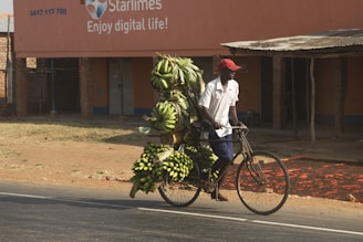 A person wearing a red cap and white shirt pushes a bicycle heavily loaded with bunches of green bananas. The background features a building with an advertisement for Starlimes, stating 'Enjoy digital life!' The setting is an outdoor urban area with a partly sunny atmosphere.