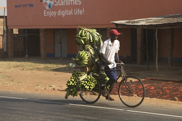 A person wearing a red cap and white shirt pushes a bicycle heavily loaded with bunches of green bananas. The background features a building with an advertisement for Starlimes, stating 'Enjoy digital life!' The setting is an outdoor urban area with a partly sunny atmosphere.