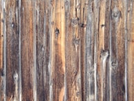 Weathered wooden planks with a rustic appearance, displaying a variety of brown and gray tones. The planks feature prominently visible wood grain and knots, enhancing the textured appearance typical of aged wood.