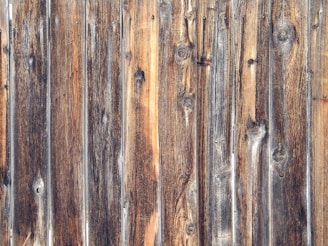 Weathered wooden planks with a rustic appearance, displaying a variety of brown and gray tones. The planks feature prominently visible wood grain and knots, enhancing the textured appearance typical of aged wood.