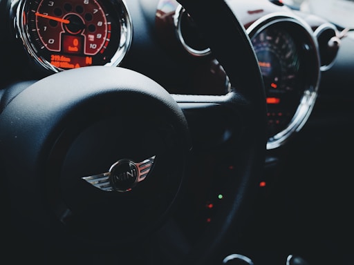 A close-up of a car interior focusing on the steering wheel and dashboard. The steering wheel has an emblem in the center with a minimalist design. The dashboard features illuminated gauges displaying speed and other metrics, highlighted by a prominent orange-red display.