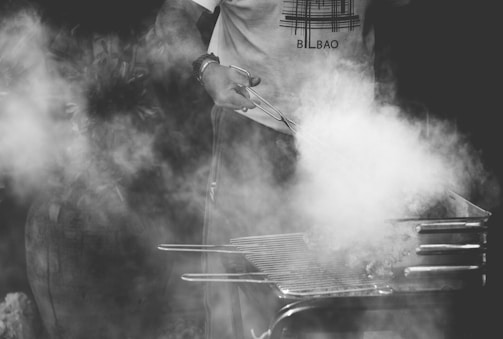 A person wearing a shirt with a design and text is cooking on a barbecue grill, surrounded by a cloud of smoke. The scene is outdoors, with a focus on the hands holding tongs and the smoky grill.