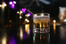 A glass of whiskey with ice and a straw sits on a dimly-lit bar counter. The background is blurred, featuring soft, colorful lights in shades of purple and yellow creating a bokeh effect.