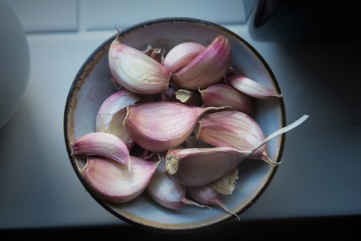 A ceramic bowl filled with several cloves of garlic, each displaying a light purple and white coloration. The bowl rests on a light-colored surface, and the composition is softly lit, creating a natural and organic feel.