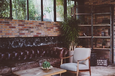 A cozy and stylish interior space featuring a brown leather chesterfield sofa set against a rustic brick wall. There is a large window allowing natural light to filter in, and a tall plant adds a touch of greenery. A modern armchair and a wooden coffee table with a small potted plant complete the seating area. Shelving units on the right hold decorative items.