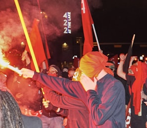 A nighttime protest with masked demonstrators holding red flags under streetlights.