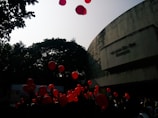 Red balloons float in the air against the backdrop of a large concrete building and trees. The scene appears to involve a crowd of people, possibly at a celebration or event.