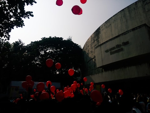 Red balloons float in the air against the backdrop of a large concrete building and trees. The scene appears to involve a crowd of people, possibly at a celebration or event.