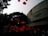 Red balloons float in the air against the backdrop of a large concrete building and trees. The scene appears to involve a crowd of people, possibly at a celebration or event.