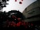 Red balloons float in the air against the backdrop of a large concrete building and trees. The scene appears to involve a crowd of people, possibly at a celebration or event.