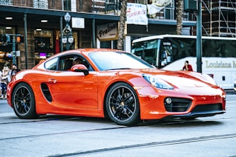 A sleek red sports car is parked on a city street, with a backdrop of urban buildings and a bus advertising tourism. The setting includes a few pedestrians and street lamps, capturing a blend of modernity and urban lifestyle.