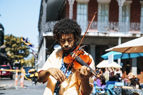A musician energetically plays a violin on a sunny street, surrounded by buildings with intricate ironwork balconies. People are sitting outdoors under large umbrellas at what appears to be a street caf&eacute;. The musician's intense focus on his craft and the vibrant street atmosphere suggest a lively urban setting.