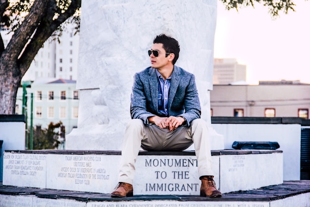 A man wearing sunglasses and a blue blazer sits on a stone monument with an inscription 'Monument to the Immigrant.' The setting appears to be urban, with buildings and trees in the background.