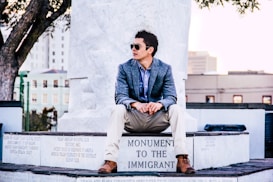 A man wearing sunglasses and a blue blazer sits on a stone monument with an inscription 'Monument to the Immigrant.' The setting appears to be urban, with buildings and trees in the background.