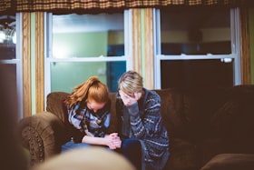 Duas mulheres sentadas em um sofá com a mão no rosto e cabeça baixa demonstrando tristeza