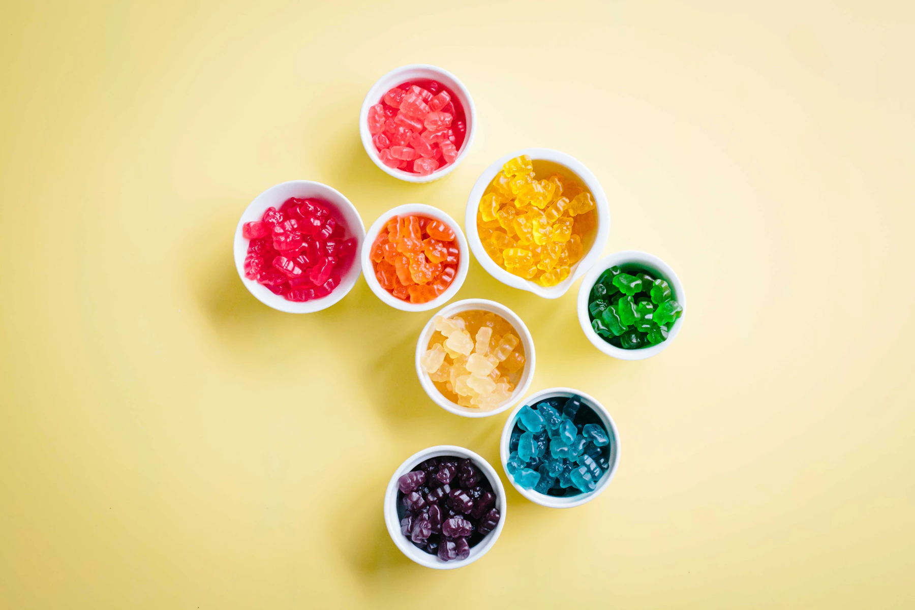 Small white bowls of gummy bears sorted by color on a pale yellow table, seen from above