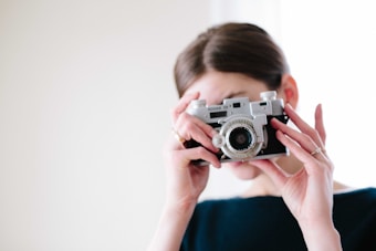 A person is holding a vintage camera to their face, capturing the moment. The background is softly blurred, highlighting the classic design of the camera with a large lens and detailed knobs.