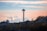 The image features the Space Needle, a distinctive tower situated in a city landscape. It is surrounded by a mix of residential and commercial buildings with visible trees. The sky has a gradient of soft pastel hues, including pink and orange, against a backdrop of white clouds.