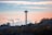 The image features the Space Needle, a distinctive tower situated in a city landscape. It is surrounded by a mix of residential and commercial buildings with visible trees. The sky has a gradient of soft pastel hues, including pink and orange, against a backdrop of white clouds.
