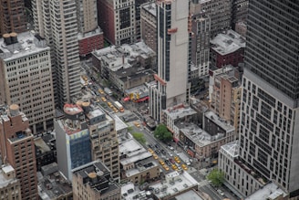 Aerial view of a densely populated urban area with numerous high-rise buildings and a busy intersection. Several yellow taxis and other vehicles are visible on the streets, along with a few areas of greenery. The buildings vary in size and design, with some under construction.