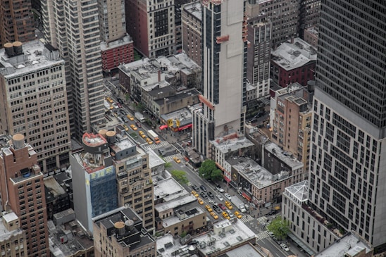 Aerial view of a densely populated urban area with numerous high-rise buildings and a busy intersection. Several yellow taxis and other vehicles are visible on the streets, along with a few areas of greenery. The buildings vary in size and design, with some under construction.