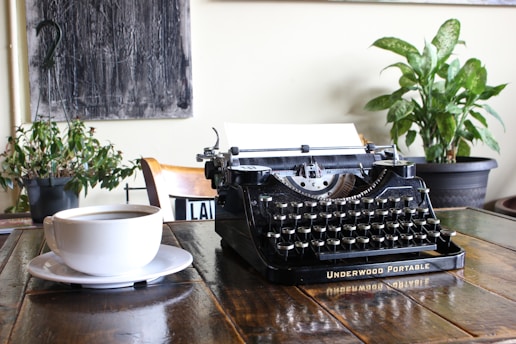 A vintage typewriter on a wooden desk surrounded by old newspapers and a steaming cup of coffee.