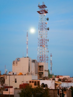 A cityscape featuring several tall communication towers against a clear blue sky. The largest tower is red and white, adorned with numerous satellite dishes and antennas. Below, industrial and residential buildings are clustered tightly together, displaying a variety of architectural styles and colors.