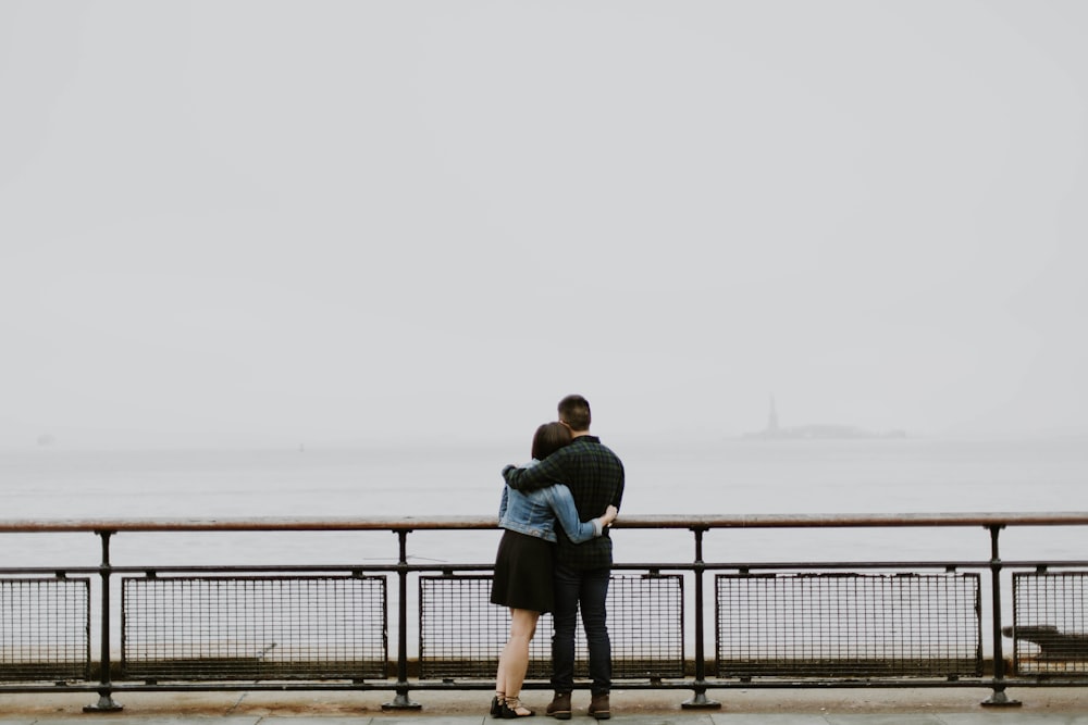A couple holds one another on a pier overlooking a body of water and overcast sky