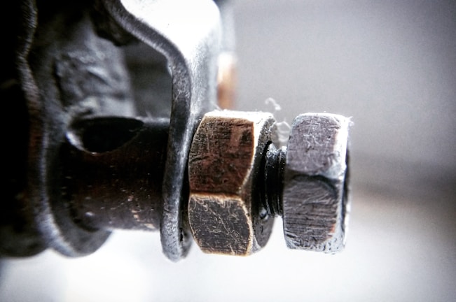 A close-up view of a metal bolt and nut, showing detailed textures and wear on the surfaces. The bolt is inserted through a hole in a metallic structure, and the background is blurred, creating a depth of field effect.