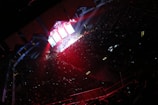 A large indoor venue filled with a crowd, illuminated by colorful stage lights. The stage is brightly lit with red and blue hues, and multiple screens display visuals. The audience is mostly in shadow, with small lights scattered throughout.