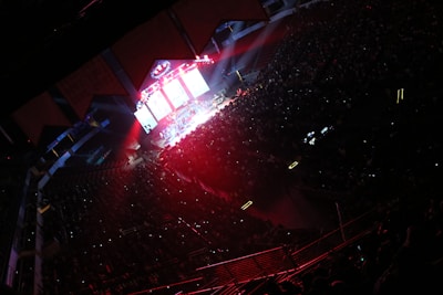 A large indoor venue filled with a crowd, illuminated by colorful stage lights. The stage is brightly lit with red and blue hues, and multiple screens display visuals. The audience is mostly in shadow, with small lights scattered throughout.