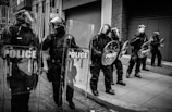 Several police officers in riot gear stand in formation on a city street. They hold shields marked 'POLICE' and are wearing helmets with visors, protective clothing, and boots. The background features urban architecture with a sign indicating a snow emergency route and no parking.