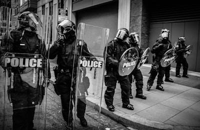 Several police officers in riot gear stand in formation on a city street. They hold shields marked 'POLICE' and are wearing helmets with visors, protective clothing, and boots. The background features urban architecture with a sign indicating a snow emergency route and no parking.