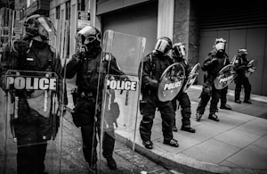 Several police officers in riot gear stand in formation on a city street. They hold shields marked 'POLICE' and are wearing helmets with visors, protective clothing, and boots. The background features urban architecture with a sign indicating a snow emergency route and no parking.