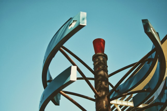A sculptural installation featuring curved metallic panels and a central cylindrical structure with a red top set against a clear blue sky.