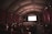 A dimly lit movie theater with rows of red cushioned seats occupied by people watching a film. The auditorium has ornate architectural details with a curved ceiling and classic wall designs. The large screen at the front is illuminated, casting a glow over the audience.