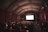 A dimly lit movie theater with rows of red cushioned seats occupied by people watching a film. The auditorium has ornate architectural details with a curved ceiling and classic wall designs. The large screen at the front is illuminated, casting a glow over the audience.