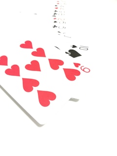 A spread of playing cards on a white background, prominently featuring the red hearts from a deck, including a close-up of the ten of hearts. The cards are arranged in an overlapping pattern with numbers and suits visible.
