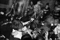 A live performance featuring a group of musicians energetically playing instruments on stage. The black-and-white image captures a crowd of enthusiastic audience members with raised arms, displaying a lively and intense atmosphere. The setting includes a vocalist, guitarist, and drummer with visible motion blur, suggesting dynamic movement.