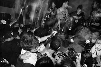 A live performance featuring a group of musicians energetically playing instruments on stage. The black-and-white image captures a crowd of enthusiastic audience members with raised arms, displaying a lively and intense atmosphere. The setting includes a vocalist, guitarist, and drummer with visible motion blur, suggesting dynamic movement.