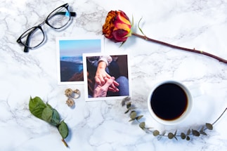 On a white marble surface, there are a pair of black-rimmed glasses, two photographs depicting scenes of nature and hand-holding, a red and yellow rose, a cup of black coffee, a pair of golden earrings, and a dried eucalyptus branch. The arrangement suggests a personal, intimate setting.