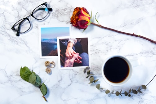 On a white marble surface, there are a pair of black-rimmed glasses, two photographs depicting scenes of nature and hand-holding, a red and yellow rose, a cup of black coffee, a pair of golden earrings, and a dried eucalyptus branch. The arrangement suggests a personal, intimate setting.