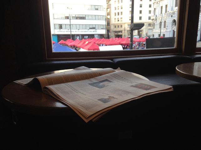 A newspaper is laid out on a round wooden table near a window. The view outside shows a row of red and blue tents in a public space, with buildings in the background. The light coming through the window creates a warm, subdued atmosphere inside.