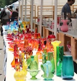 A collection of colorful glassware, including vases and decorative objects, is displayed on a long table at an outdoor market. The glass items come in a variety of vibrant colors such as red, orange, blue, green, and yellow, creating a visually striking display. Several people are browsing the market in the background.