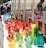 A collection of colorful glassware, including vases and decorative objects, is displayed on a long table at an outdoor market. The glass items come in a variety of vibrant colors such as red, orange, blue, green, and yellow, creating a visually striking display. Several people are browsing the market in the background.
